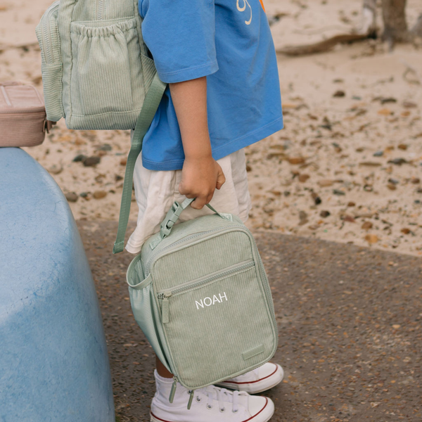 A child wearing a blue shirt and white shorts carries a sage green corduroy personalized kids lunch box with the name 'NOAH' printed in white on the front pocket, set against a beach background.